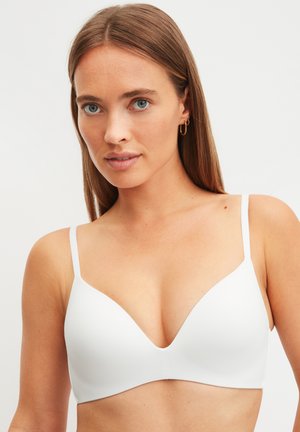 Frau mit glatten braunen Haaren, die kleine goldene Creolen und einen weißen geformten BH trägt, vor einem schlichten beigen Hintergrund.