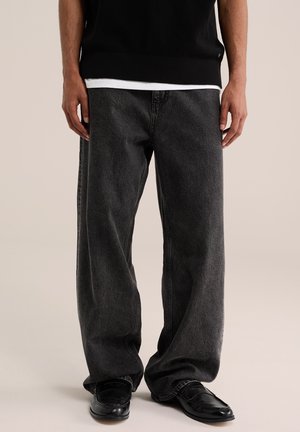 Männlich, trägt lockere dunkelgraue Denim-Jeans, schwarze Leder-Loafer und einen schwarzen Pullover mit sichtbarem weißen Hemdsaum.