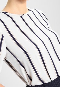 Bílý top s námořnickými a béžovými svislými pruhy, kulatým výstřihem a krátkými rukávy. Materiál má texturovaný povrch.