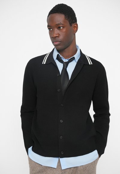 Nuori mies, joka pukeutuu mustaan neuletakkiin, jossa on valkoiset raidat kauluksessa, vaaleansiniseen paitaan, mustaan solmioon ja vaaleisiin housuihin, seisoo kädet taskuissa.