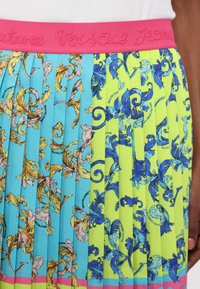 Gonna plissettata con una vita rosa. Da un lato presenta uno sfondo blu con motivi floreali gialli, mentre dall'altro lato ha una base verde neon con disegni blu.