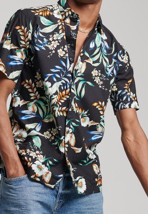 Mand iført sort kortærmet skjorte med knapper og flerfarvet tropisk blomsterprint samt lyseblå jeans, hænderne i lommerne.