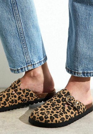 Leopard print clogs made of soft material with a black sole. Features a buckled strap and an open back design. Worn with rolled-up denim.