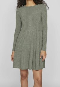 Femme portant une robe en maille côtelée vert olive à manches longues, avec un col rond et une jupe évasée.