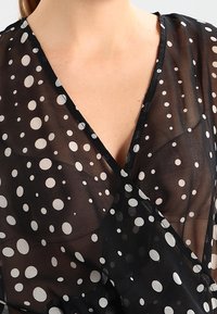 Primo piano di una persona che indossa una blusa nera trasparente a pois bianchi con scollatura a V profonda che mette in mostra la pelle chiara.
