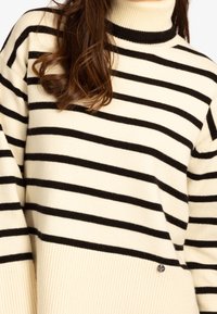Pull rayé en crème et noir, avec un col montant à nervures et un bas à nervures. Texture douce, avec un petit logo près de l'ourlet.