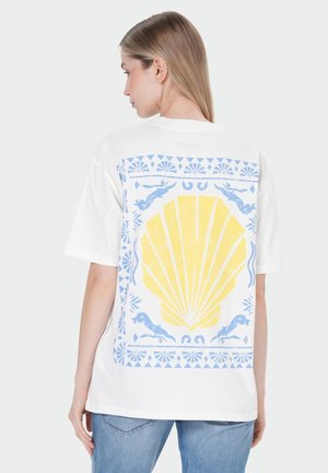 Frau trägt weißes T-Shirt mit großem gelbem Muschelmotiv und blauem dekorativem Rand auf dem Rücken, kombiniert mit hellblauen Jeans.