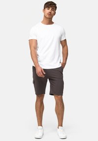 Ung mand står iført en ensfarvet hvid t-shirt, mørkegrå cargo-shorts og hvide sneakers foran en ensfarvet lys baggrund.