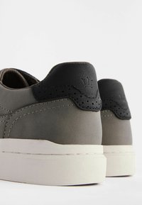 Sneakers in pelle grigia con un accento in pelle nera sul tallone. Presentano perforazioni sottili e una suola in gomma bianca.