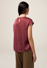 Top in satin bordeaux con maniche corte e dettaglio a chiave nella parte posteriore. Presenta una texture liscia e una vestibilità comoda. Abbinato a pantaloni marroni.