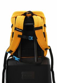 Gelber gepolsterter Rucksack mit schwarzen Riemen, Netzrücken und blauen Beschlägen; an einem schwarzen Koffer mit einem Anhängeschild befestigt.
