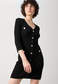 Robe en tricot noire à manches trois-quarts, décolleté en V, boutons argentés et deux poches avant. Tissu texturé avec une coupe ajustée.