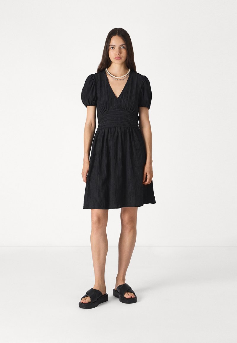 Μαύρο φόρεμα με φουσκωτά μανίκια, V-λαιμόκοψη και μαζεμένη μέση. Υφασμα με υφή, μή�κους ως το γόνατο. Μοντέλο που φοράει μαύρες σανδάλια και πέρλα κολιέ.