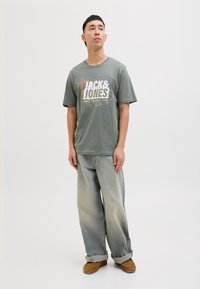 Tamsiai žalios spalvos medvilnės marškinėliai su balta, oranžine ir geltona "JACK & JONES" grafika. Suderinti su šviesiai mėlynais džinsais ir rudomis šlepetėmis.