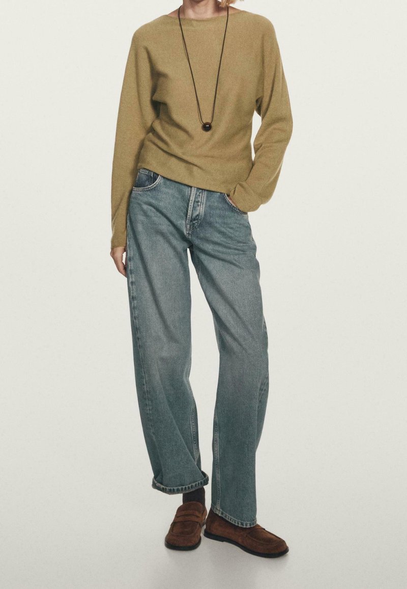 Personne portant un pull à manches longues couleur marron clair, un jean bleu ample, des mocassins marron et un long collier avec un pendentif rond, debout avec une main dans la poche.