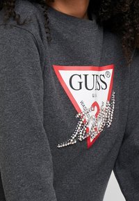 Sudadera gris con un gran logo de triángulo rojo que incluye el texto "GUESS", adornada con detalles de strass para un toque adicional.