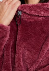 Mano ajustando el cordón negro en el cuello de una chaqueta de forro polar de textura suave y color rojo oscuro.