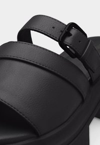 Sandale en cuir noir avec larges brides et fermeture à boucle ajustable sur une semelle épaisse, en gros plan.