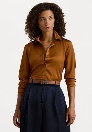 Camicia in raso marrone con colletto classico e maniche lunghe, abbinata a pantaloni a gamba larga blu scuro e una cintura marrone con fibbia argentata.