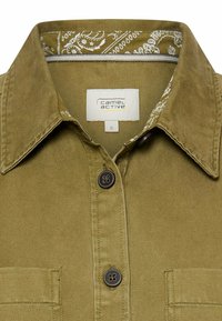 Oliwkowa koszula zapinana na guziki z kołnierzem wyściełanym materiałem w wzór bandany, z dwoma kieszeniami z przodu i brązowymi guzikami. Widoczna metka z rozmiarem.