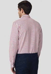 Mężczyzna odwrócony tyłem, ubrany w biały-shirt w pionowe czerwone paski i granatowe spodnie na tle jednolitego jasnego tła.