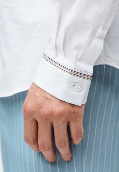 Άσπρο μανίκι πουκάμισου με πολύχρωμη ριγέ ραφή, με ένα κουμπί και λείο ύφασμα. Το χέρι είναι μερικώς ορατό, φοράει το μανίκι.