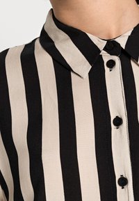 Czarna i beżowa koszula w paski z kołnierzykiem, z przodem zapinanym na guziki i czarnymi guzikami. Materiał wydaje się gładki z lekko połyskującą powierzchnią.