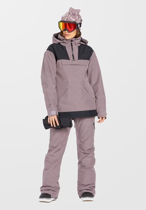 Snowboardoutfit met een lichtpaarse anorak met een zwart paneel, losse lichtpaarse broeken en een gemusterde paarse beanie.