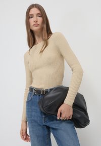 Beige Rippenoberteil mit langen Ärmeln und figurbetonendem Design, kombiniert mit hochtaillierten blauen Jeans. Hält eine große schwarze Lederhandtasche.