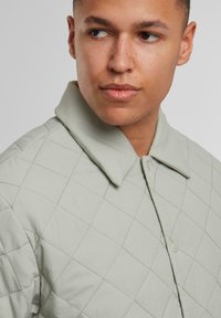 Hellgrüne gesteppte Jacke mit einem breiten Kragen, die über ein Rautenmuster und einen einzelnen Druckknopfverschluss verfügt. Glattes, strukturiertes Material.