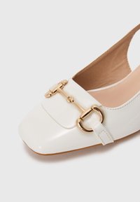 Valkoinen patentoitu nahkainen tasapohjainen kenkä, jossa on neliön muotoinen kärki ja kultainen metallinen hevosenpäädytön yksityiskohta edessä, beige sisustus ja ruskea pohja.