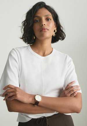 Selvsikker kvinde med kort krøllet hår iført en hvid skjorte, guldringeøreringe, et armbåndsur og mørke bukser med snøre.