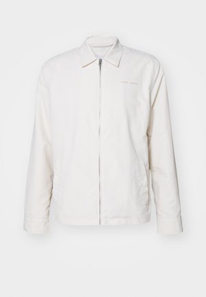 Bela jopica na zadrgo iz mehke tkanine. Ima ostri ovratnik, dolge rokave in subtilno blagovno oznako na prsih.