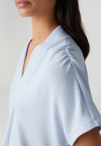 Haut bleu clair à col en V avec un tissu texturé, une coupe douce et des manches courtes. Présente des accents froncés au niveau des épaules.