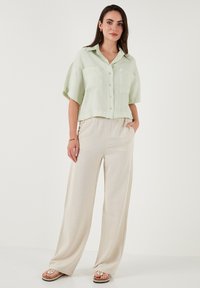 Camicia verde chiaro a maniche corte con bottoni e due tasche sul petto, abbinata a pantaloni beige a gamba larga, indossata con sandali di colore chiaro.