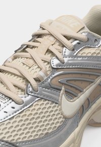Μπεζ και ασημί παπούτσι τρεξίματος Nike με άνω μέρος από δίχτυ, κρέμ κορδόνια και υφασμένο λογότυπο Nike swoosh στο πλευρό.