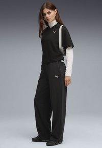 Young woman wearing black Puma tracksuit, white long-sleeve top underneath, black shoes, and carrying a pale shoulder bag against gray background.