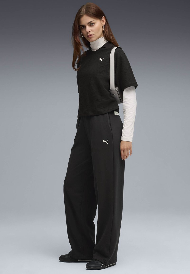 Young woman wearing black Puma tracksuit, white long-sleeve top underneath, black shoes, and carrying a pale shoulder bag against gray background.