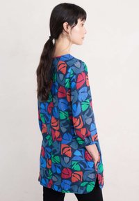 Tunic a maniche lunghe realizzata in un tessuto blu con motivi di foglie colorate in rosso, verde e arancione; tasche laterali e vestibilità comoda.