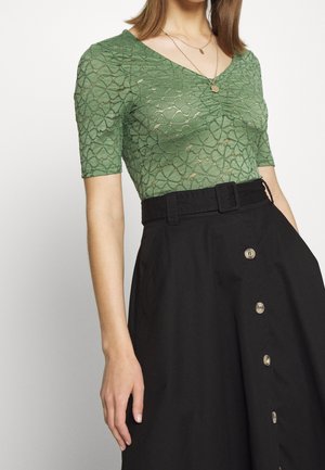 Femme portant un haut vert en dentelle à manches courtes avec un devant froncé, des colliers superposés en or et une jupe noire taille haute avec des boutons sur le devant et une ceinture.