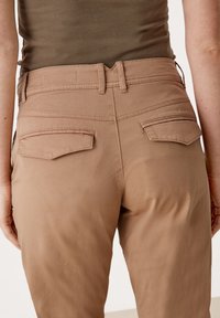 Bruna, figurnära byxor med en slät textur, som har två bakfickor och subtila sömdetaljer, sedd från baksidan.