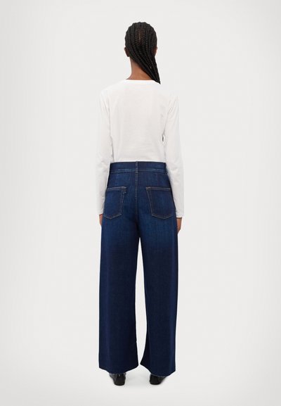 Jeans de denim de pierna ancha en azul oscuro, con cintura alta, bolsillos traseros y un ajuste holgado. Combinados con una camisa blanca de manga larga.