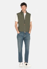 Uomo in piedi con le mani in tasca, che indossa un gilet verde senza maniche con zip, una t-shirt bianca, jeans blu e sneaker bianche con strisce nere.