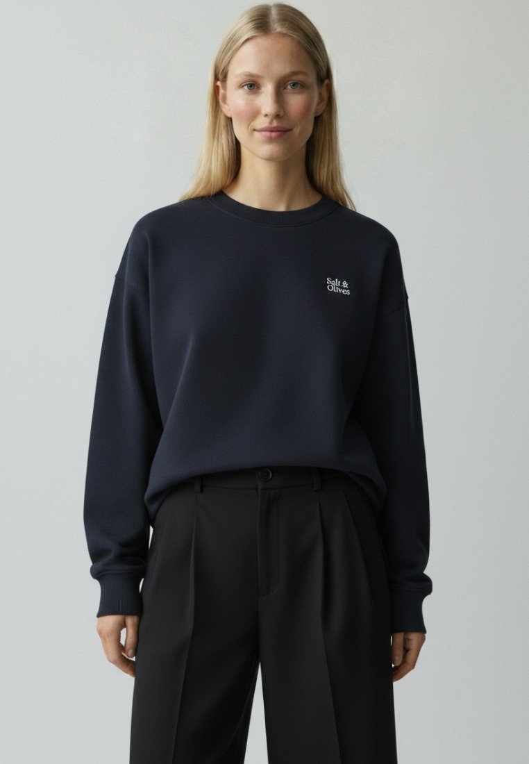 Kvinde med langt blondt hår iført en marineblå sweatshirt med "Salt & Olives"-logo og sorte plisserede bukser mod en ensartet grå baggrund.