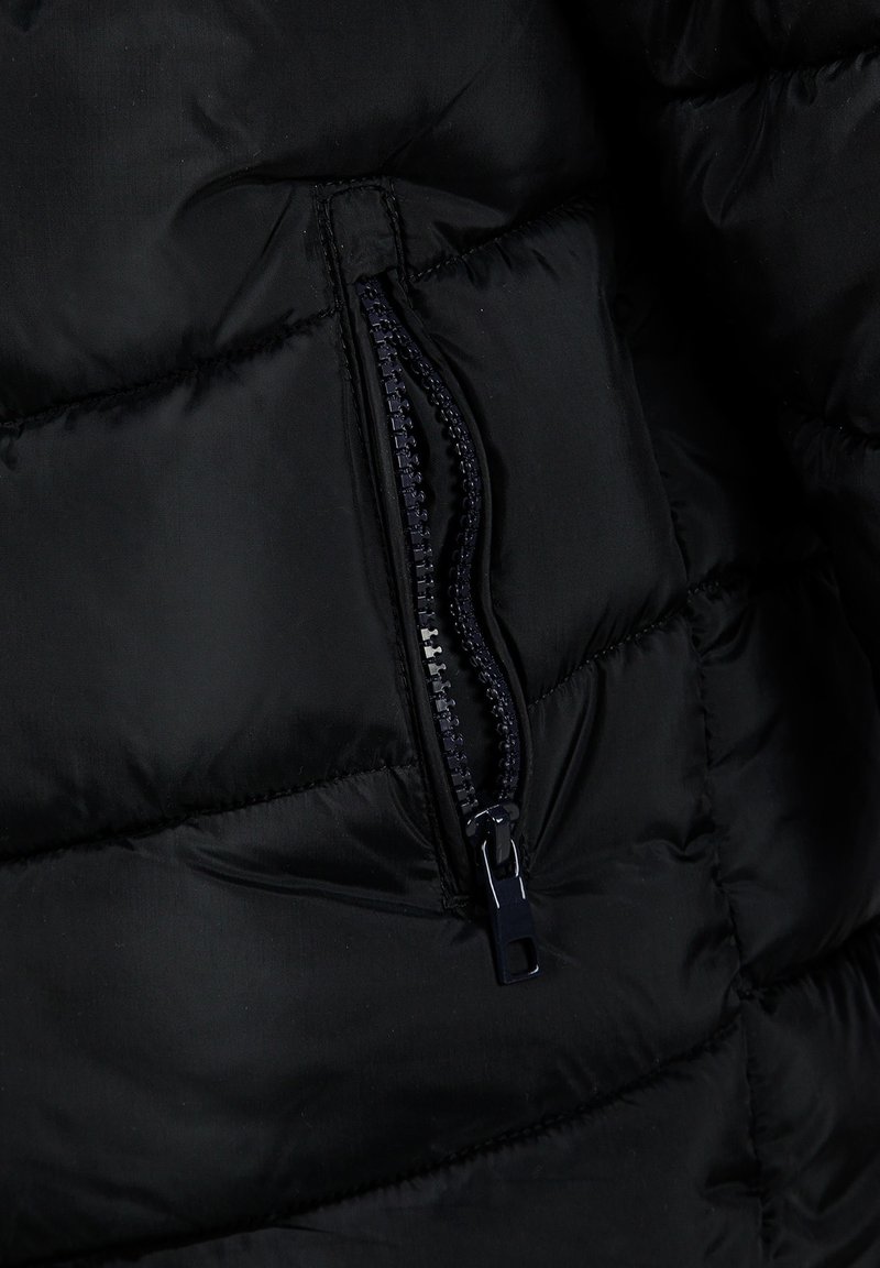 Veste rembourrée noire en tissu avec une poche zippée. La texture est lisse avec des sections matelassées et un tirette en plastique.