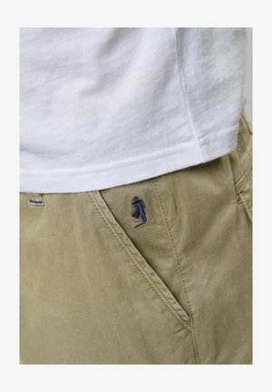 Beżowe szorty wykonane z bawełny o subtelnej strukturze, z haftowaną ciemnoniebieską postacią na przedniej kieszeni. Biała koszulka pod spodem.