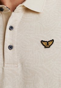 Polo créme avec un motif texturé, trois boutons foncés et un logo brodé noir et or sur la poitrine gauche.