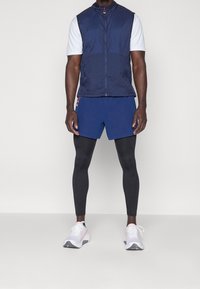 Blauer Weste, weißes Kurzarmhemd, marineblaue Shorts, schwarze Leggings und weiße Schuhe mit pinken Akzenten. Leichte Materialien mit einem taillierten Design.