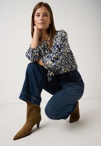 Bluse mit Blumenmuster in Marineblau mit gelben und weißen Blumen, kombiniert mit weit geschnittenen Denim-Jeans. Sandfarbene, spitze Wildleder-Stiefeletten.