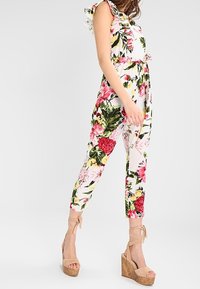 Floral jumpsuit with ruffled sleeves, featuring vibrant pink and green patterns, fitted waist, and tapered legs; paired with nude platform sandals.